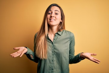 Young beautiful blonde woman with blue eyes wearing green shirt over yellow background clueless and confused expression with arms and hands raised. Doubt concept.