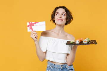 Young woman girl in casual clothes hold in hand gift certificate makizushi sushi roll served on plate traditional japanese food isolated on yellow background studio portrait. People lifestyle concept