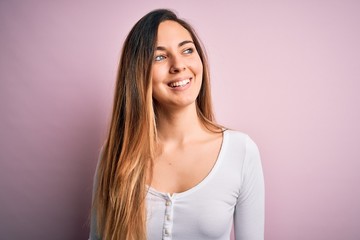 Obraz premium Young beautiful blonde woman with blue eyes wearing white t-shirt over pink background looking away to side with smile on face, natural expression. Laughing confident.