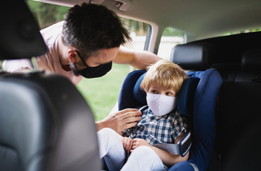 Father putting small son in car seat before trip, wearing face masks.
