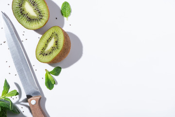 top view of tasty kiwi fruits near green peppermint, knife and black seeds on white