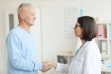 Obraz premium Waist up side view portrait of smiling female doctor shaking hands with senior patient while standing in clinic interior