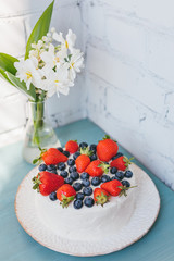 Wonderful summer cake with strawberry, blueberry and bouquet of narcissus flowers with lily of the valley in a vase on the blue table. Copy space