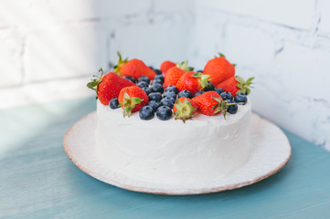 Close-up of delicious home made celebratory summer cake with whipped cream frosting and berries decoration on blue wooden table. Bluberry and strawberry