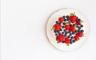 A top view of home made cake or pie on beautiful white handmade plate on light wooden background with copy space. Dessert is decorated with summer berries, strawberries and blueberries