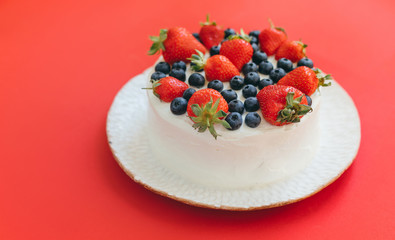 A tasty summer pie or cake with strawberries and blueberries on white handmade plate on juicy red background. Delicious dessert with berries for celebration