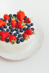 A cut view of home made cream cake on beautiful white handmade plate on light wooden background. Dessert is decorated with summer berries, strawberry and blueberry