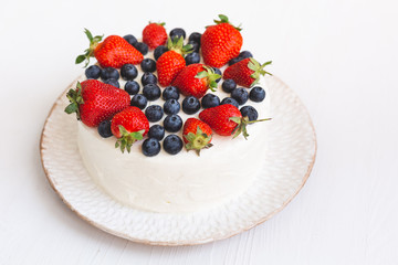 A beautiful appetizing cake with white cream frosting and fresh summer berries on hand made plate on white wooden background. Delicious home made dessert with blueberries and strawberries