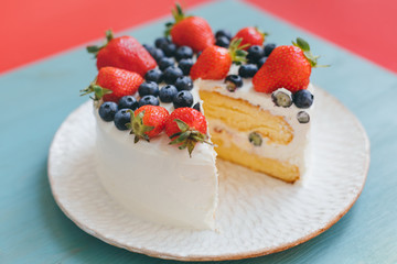 Tasty sweet home made cut birhday cake on interesting handmade plate on blue and red background. A dessert is decorated with fresh blueberries and strawberries. Summer mood