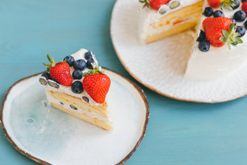 Close-up of piece of birthday tasty sweet sponge cake with white whipped cream and summer berries. Dessert decorated with fresh juicy blueberries and strawberries. Light blue background