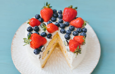 A wonderful cut sponge cake with whipped cream, blueberries and strawaberries on light blue background. Sweet home made dessert for celebration. Selective focus