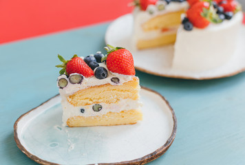 A piece of tasty sponge cake with white whipped cream, fresh juicy blueberries and strawaberries on beautiful handmade plates on blue and red background. Sweet summer home made dessert for birthday 
