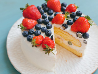 Beautiful delicious cut sponge cake with whipped cream and fresh summer berries on handmade plate on light blue background. Home made dessert with blueberries and strawberries. Selective focus