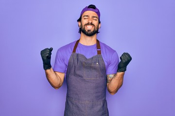 Young handsome hispanic man with bear wearing professional apron working as tattoo artist very happy and excited doing winner gesture with arms raised, smiling and screaming for success. Celebration