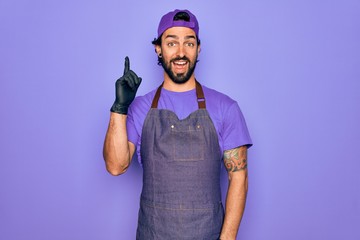 Young handsome hispanic man with bear wearing professional apron working as tattoo artist pointing finger up with successful idea. Exited and happy. Number one. - Powered by Adobe