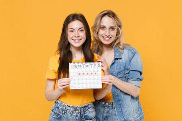 Smiling young women girls friends in denim clothes posing isolated on yellow background studio. Medical, healthcare, gynecological concept. Hold female periods calendar for checking menstruation days.