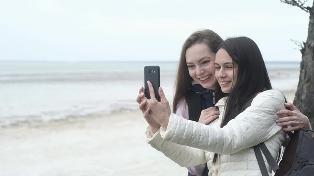 two young girls with backpacks on backs are standing at the edge of the sea cliff. Girls travel to sea and take Funny selfies in nature with their phone camera. Active walks along the coast together