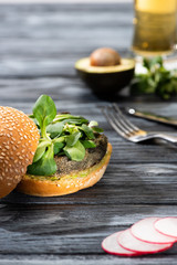selective focus of tasty vegan burger with microgreens served on wooden table with radish, avocado half, cutlery and beer