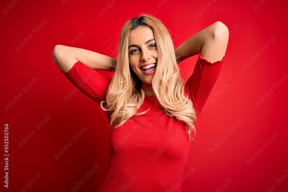 Poster Young beautiful blonde woman wearing casual t-shirt standing over isolated red background relaxing and stretching, arms and hands behind head and neck smiling happy