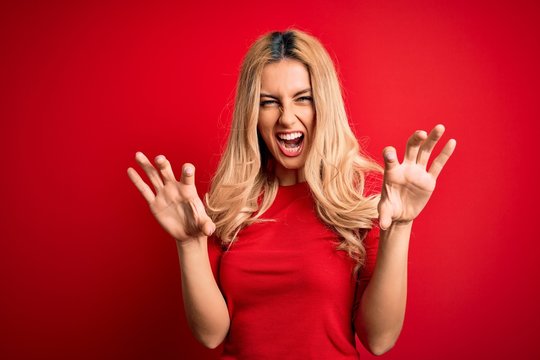 Young beautiful blonde woman wearing casual t-shirt standing over isolated red background smiling funny doing claw gesture as cat, aggressive and sexy expression