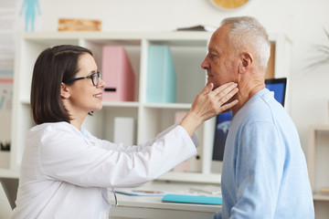 Obraz premium Side view portrait of smiling female doctor palpating neck of senior patient while examining him during consultation in clinic