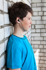 Preteen Caucasian boy listening music with earphones, standing close to a white brick wall, side view
