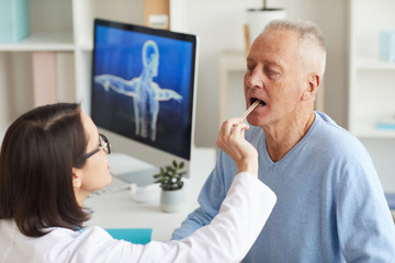 High angle view at female doctor checking throat of senior patient while examining him during consultation in clinic