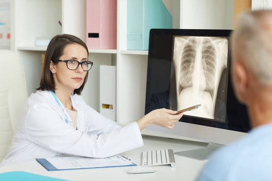 Portrait of serious female doctor pointing at x-ray image of lungs and chest while consulting senior patient in clinic