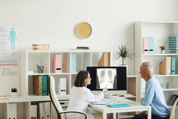 Wide angle side view at female doctor pointing at x-ray image of lungs and chest while consulting senior patient in clinic, copy space