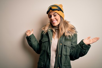 Young brunette skier woman wearing snow clothes and ski goggles over white background smiling showing both hands open palms, presenting and advertising comparison and balance