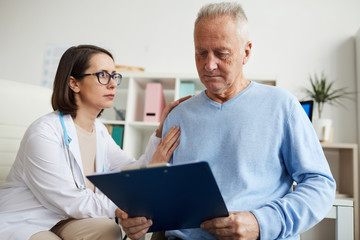 Obraz premium Portrait of caring female doctor comforting senior patient reading diagnosis on clipboard during consultation in clinic, copy space