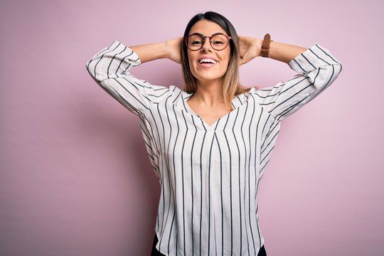 Young beautiful woman wearing casual striped t-shirt and glasses over pink background relaxing and stretching, arms and hands behind head and neck smiling happy
