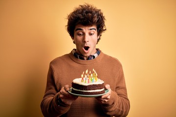 Young handsome man holding birthday cake standing over isolated yellow background scared in shock...