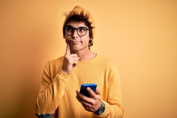 Young handsome man using smartphone wearing glasses over isolated yellow background serious face thinking about question, very confused idea