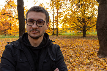 A young short-haired guy wearing glasses and a black jacket stands with his arms crossed in the autumn park. Meditation in the falling park.