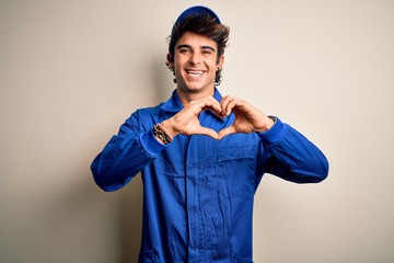 Young mechanic man wearing blue cap and uniform standing over isolated white background smiling in love doing heart symbol shape with hands. Romantic concept.