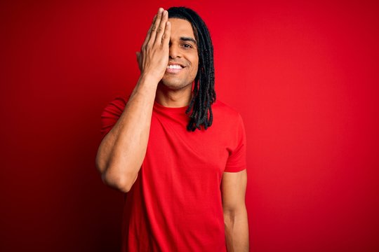 Young handsome african american afro man with dreadlocks wearing red casual t-shirt covering one eye with hand, confident smile on face and surprise emotion.