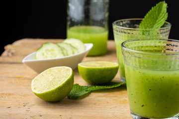 Close-up of two glasses with cucumber juice, cut limes, mint, cucumber slices and bottle, with selective focus, on wooden table, black background, horizontal