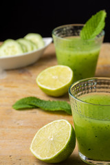 Aerial view of glasses with cucumber juice, cut limes, mint and cucumber slices, with selective focus, on wooden table, black background, vertical