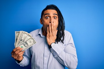 Young african american afro man with dreadlocks holding dollars over blue background cover mouth with hand shocked with shame for mistake, expression of fear, scared in silence, secret concept
