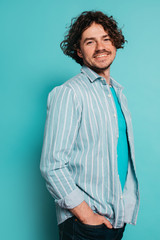 Portrait of handsome cheerful young man holding hands in pockets and posing on camera. Positive guy smiling in side view. Isolated over blue background.