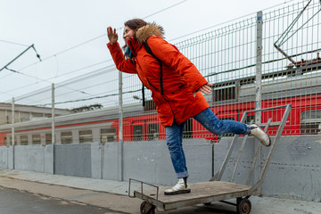 young woman in a red jacket stands on one leg on a trolley and looks into the distance