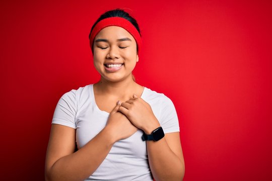 Young beautiful asian sporty woman wearing sportswear doing sport over red background smiling with hands on chest with closed eyes and grateful gesture on face. Health concept.