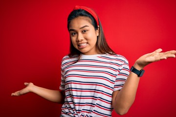 Young beautiful asian girl wearing casual striped t-shirt over isolated red background clueless and confused expression with arms and hands raised. Doubt concept.