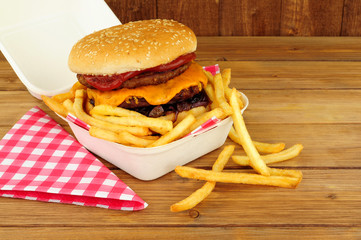 Double cheeseburger in a sesame seed covered bread bun with tomato sauce and French fries in a take away box on a wood background