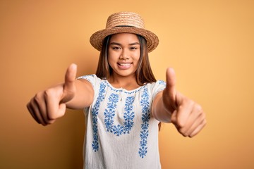 Young beautiful asian girl wearing casual t-shirt and hat standing over yellow background approving doing positive gesture with hand, thumbs up smiling and happy for success. Winner gesture.