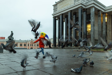 A young woman wearing red jacket and a mask (bird's head) running among a flock of pigeons in the square by the Russian state library.