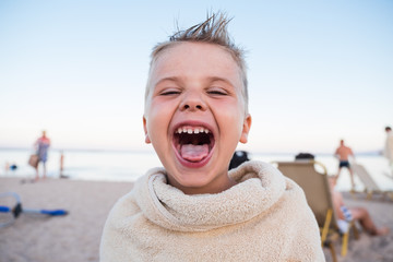 Cheerful cute and emotional preschool baby boy shows grimaces, laughs at the camera. Child is wrapped in a towel after swimming on the beach. Vacation at sea.