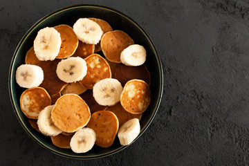 Trendy food - Tiny pancake cereal with banana in a bowl on a grey background, small pancakes, flat lay, copy space, top view. 