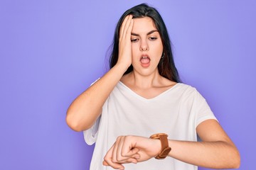 Young beautiful brunette woman wearing casual white t-shirt over purple background Looking at the watch time worried, afraid of getting late